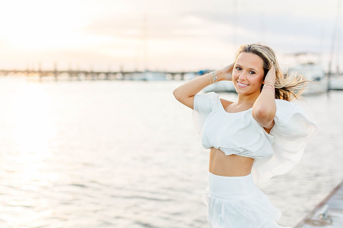 Senior girl in a white crop top and skirt smiling at sunset on the water during her coastal senior session at New Bern marina in North Carolina